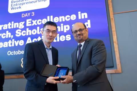 Two men in dark suit jackets hold a glass award while posing in front of a purple screen with the word "excellence" partially visible above their heads. 