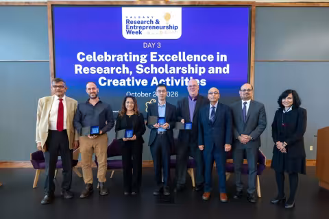Eight smiling people standing shoulder to shoulder in front of a purple screen with the text: "UAlbany Research and Entrepreneurship Week" and "Celebrating Excellence in Research, Scholarship, and Creative Activities"
