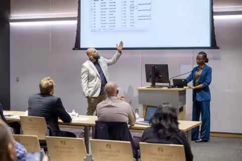 A man in a light suit jacket looks up and gestures to numbers on a screen while a woman a blue suit stands near by at a computer on a lectern.