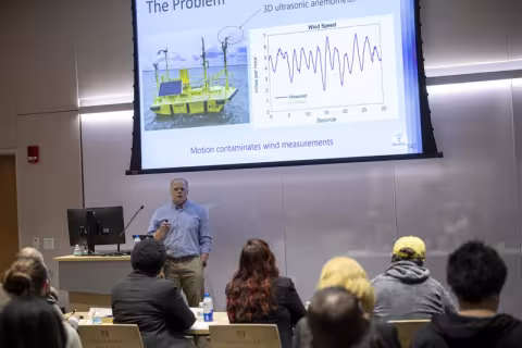 A man in a blue-collared shirt stands holding a slideshow clicker in front of an audience with a picture of floating scientific instrument and line graph on a screen behind him.