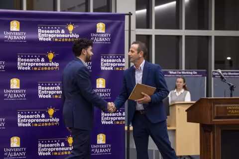 Two men shaking hands on stage in front of a purple backdrop with the words "Research and Entrepreneurship Week" on it. 