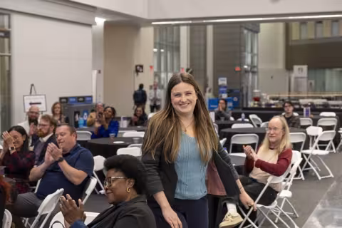 A smiling woman in a black suit and blue shirt stands as people seated around her at tables applaud.