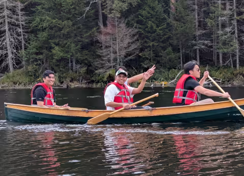Four students smile while rowing a canoe in a lake.