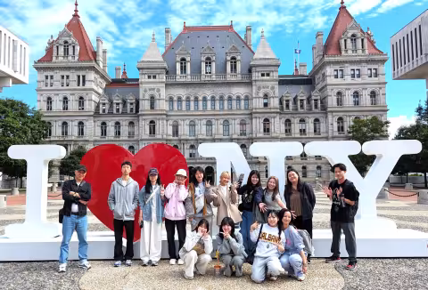 Fourteen students pose in front of an I Heart New York sculpture near the New York State Capitol Building.