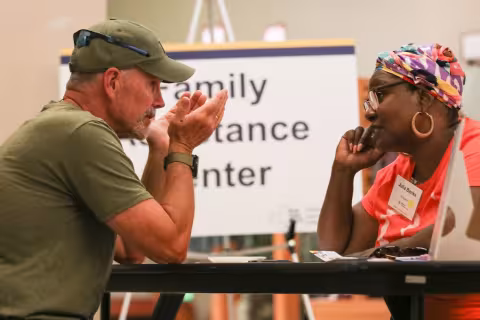 A man wearing an olive T-shirt and olive cap sits at a table speaking with a woman wearing a bright orange T-shirt and a colorful headscarf. A sign reading "Family Assistance Center" is behind them.