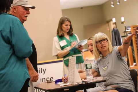 A woman seated at a table, wearing glasses and a gray T-shirt, gestures across the room as a man in a cap accepts her directions. A woman wearing a green vest writes on a clipboard in the background.