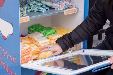 The image zooms in on the contents of the period pantry including pads and tampons, stacked on two shelves. Claire Jennings reaches in to stock the pantry. 