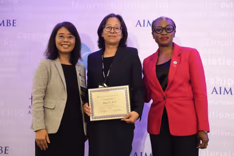 Three women smile for a photo. They are standing against a purple-lit backdrop printed with the name “AIMBE” (in purple) as well as a collection of science-related words in very faint print. The woman at left is wearing rimless glasses, a tan blazer and a black dress. The woman in the center is wearing a black blazer and black square rimmed glasses and is holding a certificate. The woman at right is wearing a red blazer and glasses with black circular rims. 