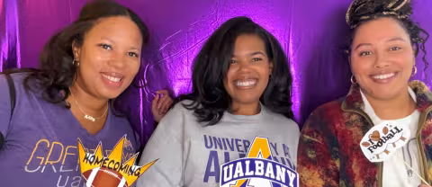 Three UAlbany alumni wearing purple and gold pose for a picture inside a photo booth.
