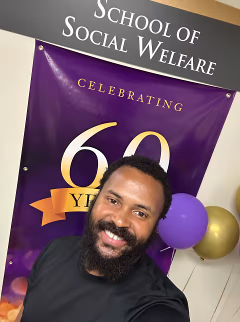 A young man with dark hair and a beard, wearing a black T-shirt, smiles in front of a purple banner that says "Celebrating 60 years". A sign reading "School of Social Welfare" hangs above the banner. 