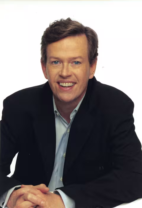 actor with dark hair smiles while wearing a white shirt and dark suit coat