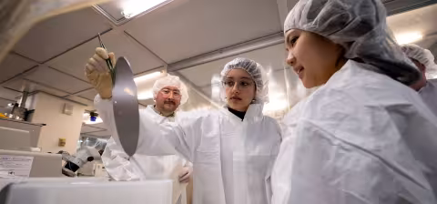 Two students in white bunny suits, hairnets and gloves work inside a clean room while their professor looks on.