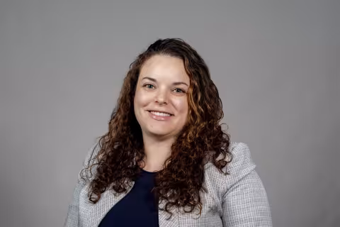 A woman with curly brown hair wearing a gray blazer and navy shirt smiles for a portrait in front of a gray backdrop.