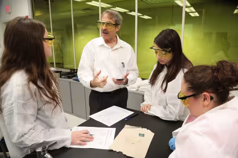 Dr. Robert Brainard wears protective glasses and gestures as he speaks to three students inside his research lab. All three students wear lab coats and protective glasses, and one takes notes.