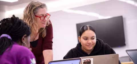 An instructor leans on a table to look at a student's laptop screen. A second student is seated across the table.