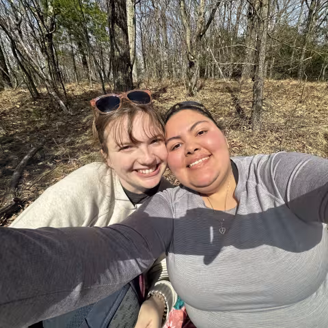 Two young women stand together, smiling for a selfie, in a forest in early spring. 