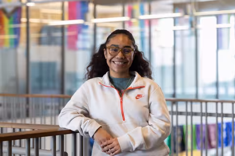 A young woman with long black hair and glasses wears a white Nike sweater and poses for a portrait on the UAlbany campus.