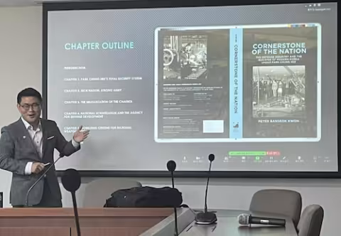 A man in a suit presents in front of a screen that shows the jacket for a book titled, "Cornerstone of the Nation"