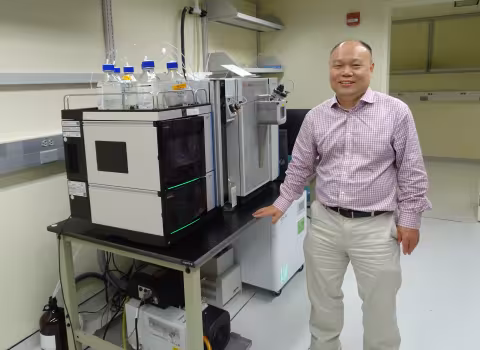 A man wearing khaki pants and a plaid button-down shirt stands, smiling, in a brightly lit lab next to a table bearing two pieces of lab equipment. Both machines are made of metal and shaped like cubes. One has a rack containing bottles of clear liquid on its top and a digital panel on the front.