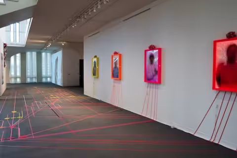 Flourescent gaffers tape create a path on the University Art Museum floor. Several pieces of tape travel up a white wall and lead to three lightbox portraits of individuals covered in text.