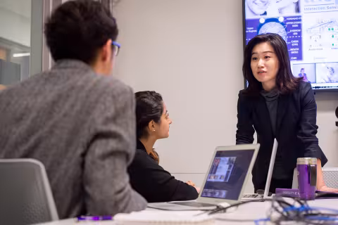 A faculty member speaks to two students with their laptops during a presentation