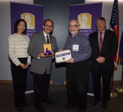 Four people stand in front of UAlbany banners, one holding a purple great Dane bust and one holding a certificate with his name, Richard Albagli, printed on it.