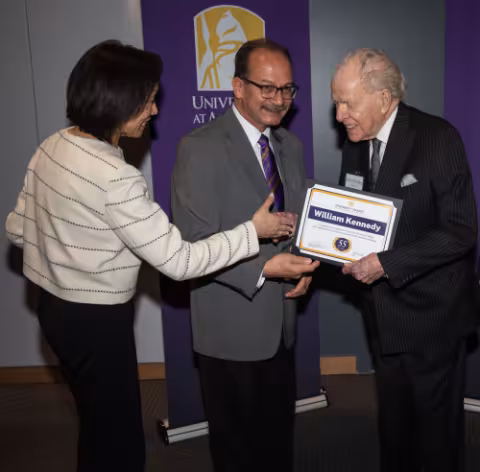 William Kennedy holds a certificate with his name printed on it, while being congratulated by Provost Carol Kim and President Havidán Rodríguez.