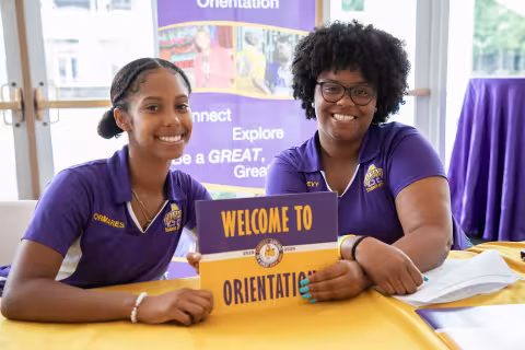 Two Orientation Leaders pose for a photo at a welcome desk, while holding a "Welcome to Orientation" sign.