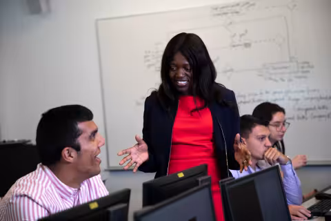 An instructor gestures and smiles as she speaks to a student seated at a desktop computer.