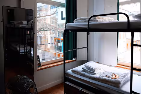 A college student's living space. A mirror sits against the wall near a bunk bed with folded clothes and sheets on the mattress.