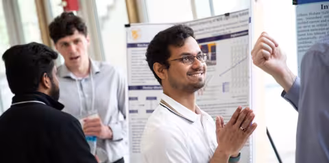A student standing in front of a research poster smiles and puts his palms together as he listens to someone off camera at UAlbany Showcase.