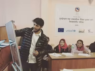 A man writes on a white board while two women are seen in the background watching