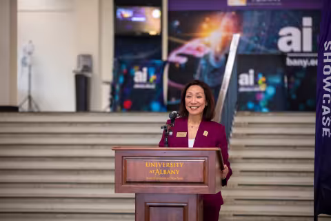 UAlbany Provost Carol Kim smiles while speaking at a University podium, with posters for the AI-Plus initiative behind her.