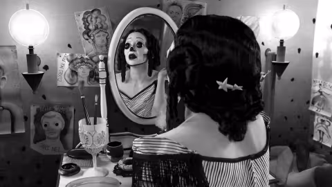 A black and white video still of the artist in costume, theatrically looking in a mirror at a vanity in a room heavily decorated with drawings.  