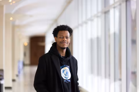 A young man with short black hair wears a black hoodie and poses for a portrait on the UAlbany campus.