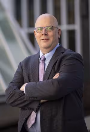 a man in glasses and a gray suit stands, arms crossed