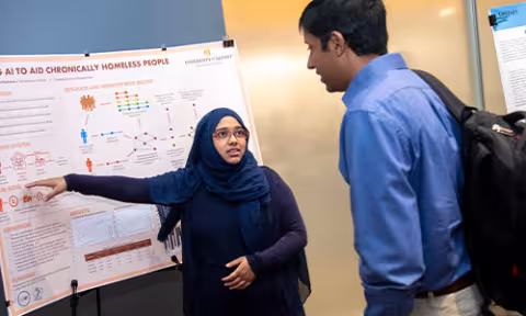 A student wearing a deep turquoise head covering, a purple sweater and glasses presents their work to a man in a blue button up shirt
