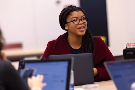 A young woman in a red blazer and black glasses works at a laptop and looks into the distance