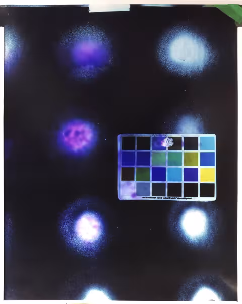 A photographic print of a gridded color swatch paper on a dark purple background, with seven dots on the surface, seemingly resulting from a chemical process.   