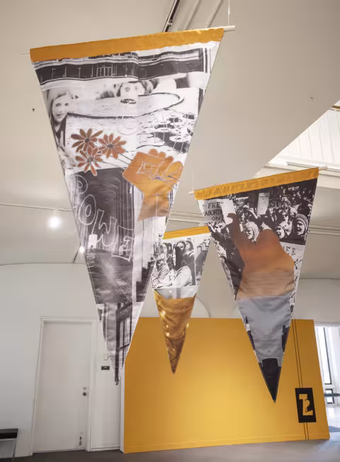  Three flag shaped banners, hanging from the ceiling, depicting collaged protest photographs.   