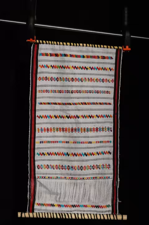 A red and white weaving with multicolored embellishments on a small loom.