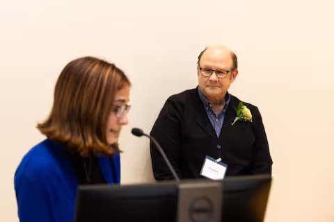 A man with glasses wears a flower lapel pinned to a sweater and leans against a wall as a student speaks into a microphone