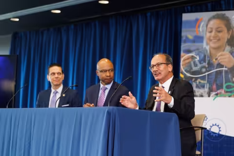 Three men in suits are seated at a table in front of microphones. One of them is speaking and gesturing.