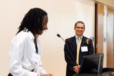 A man in a dark suit jacket and yellow tie smiles as a student speaks into a microphone nearby
