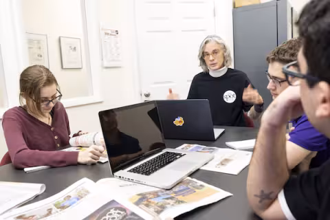 A woman with short gray hair talks to students seated at a table topped with laptops and newspapers.