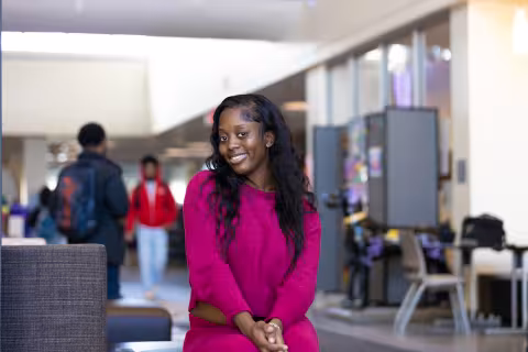 A young woman with long black hair wears a bright pink sweater and bright pink pants and poses for a portrait on the UAlbany campus