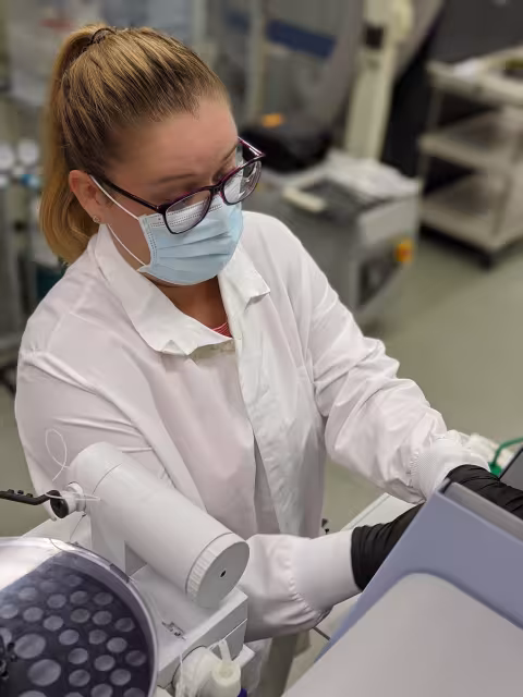 Emily wears a lab coat and a mask and sits at a desk in the Wadsworth Center.