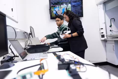 Two researchers work inside a UAlbany lab. One sits in front of a computer and points at the monitor, while the other looks on and holds a virtual reality headset.