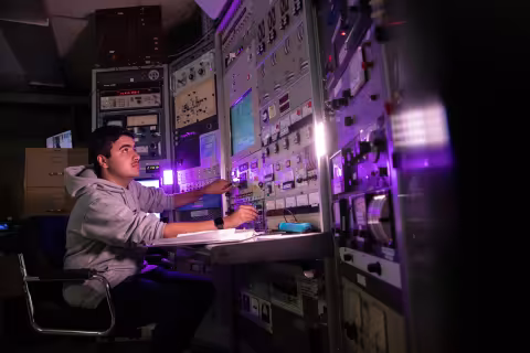 A student sits at a desk in front of a large instrument board and turns a different dial with each hand, as he looks up at the monitor.