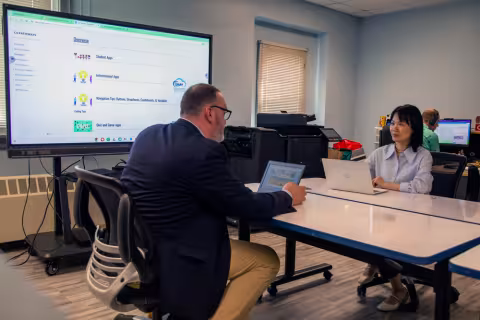 Two educators sitting at a table with laptops and a projection screen.